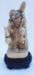 Grupo Escultórico Oriental em Marfinite, representando camponês, acompanha peanha em madeira nobre, medindo 16 cm de altura.