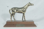 Troféu - Melhor Páreo -84- Castel, escultura representando cavalo em metal dourado sobre base de madeira, com placa de identificação. Med. 22cm de alt x27 cm de lar x l11cm de prof.