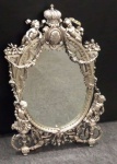 Imponente espelho em prata contrastada, decorado com querubins e guirlandas, encimado por medalhão e coroa. Medidas 50 x 30 cm.